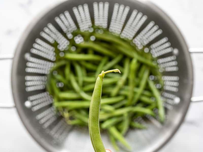 Close up of a green bean with stem, more green beans in a colander in the background
